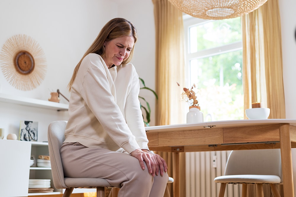 Frau sitzt am Esstisch und hält schmerzbedingt das Knie/Hüfte nach Hüft-OP